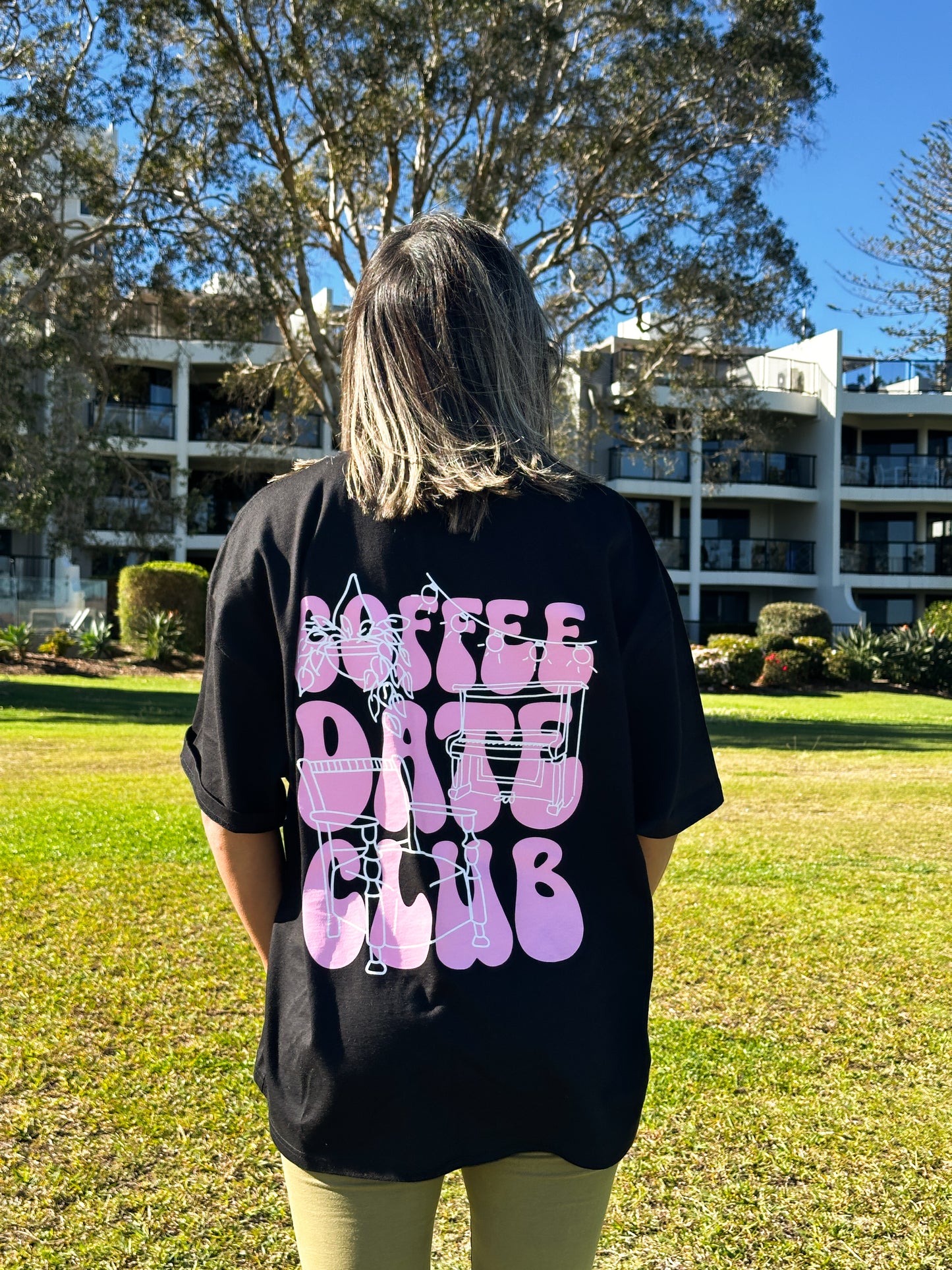 Person wearing a black oversized t-shirt with pink "Coffee Date Club" text on the back, standing in a sunny park. Gun Cotton Coffee merch.